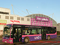 Uno bus with a purple livery in front of The Galleria shopping complex in the evening